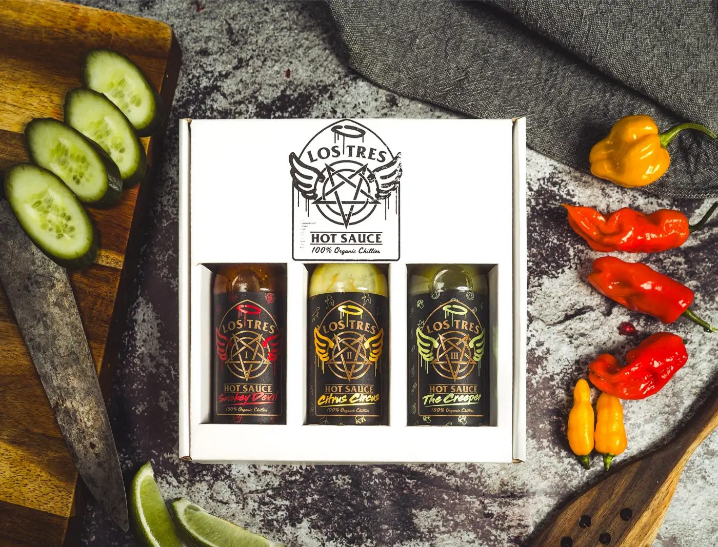 three hot sauce bottles in a white display box on stone with chillies and cucumber